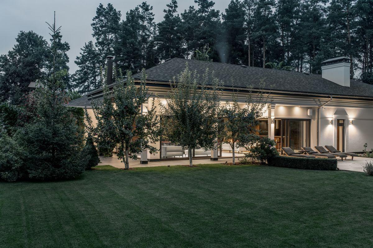 Luminous modern cottage with light walls, columns and a gray roof with chimneys on the background of the evening sky and pine trees. It has a large lawn with fruit trees, terrace with deck chairs.